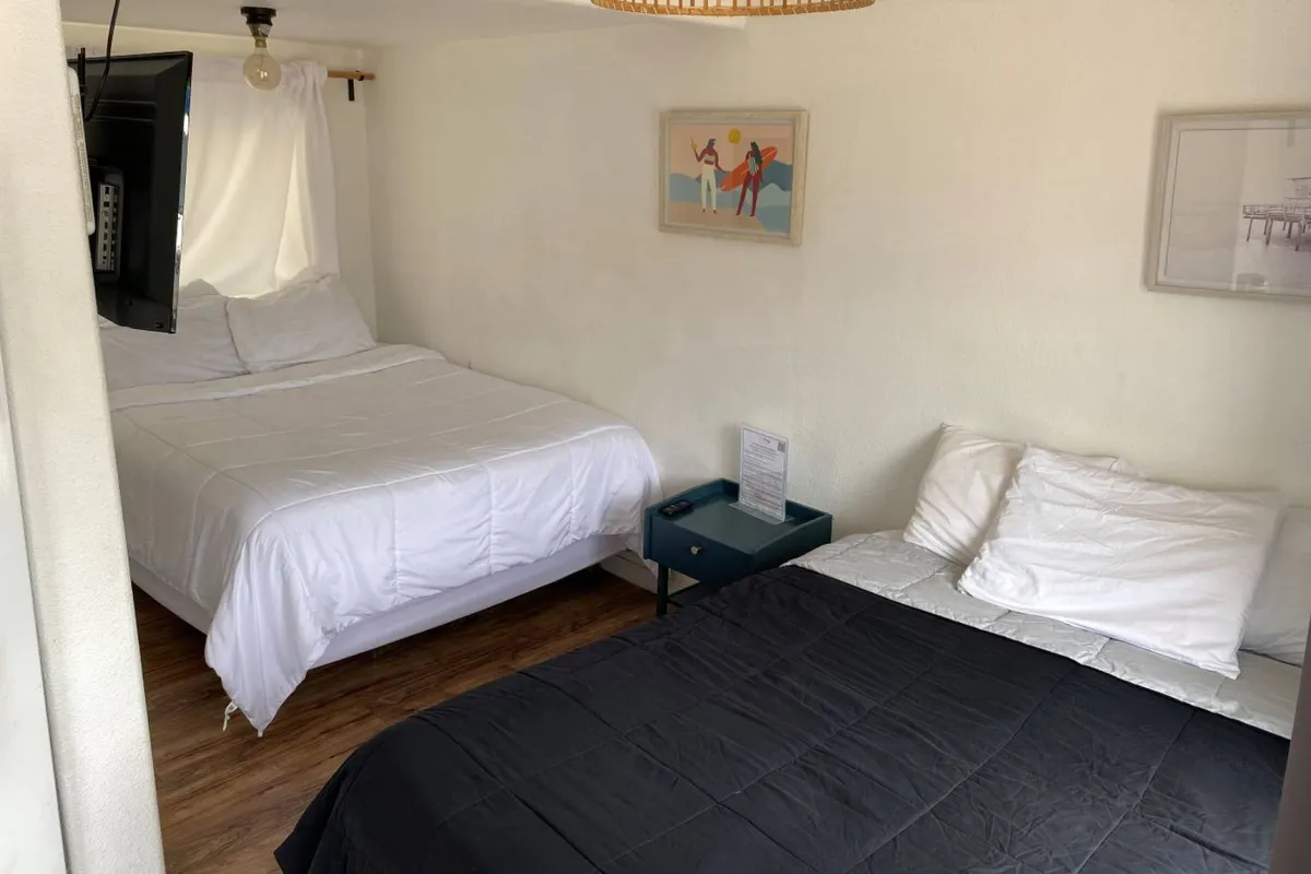 A peaceful corner of the studio features two beds with fresh, decorative bedding, a nightstand, and wall art. Natural light pours in from a nearby window, creating a restful and inviting mood ideal for unwinding after a day at the beach.
