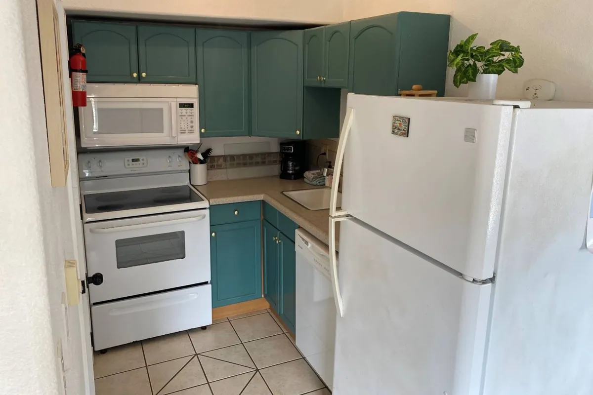 This interior shot captures a cozy kitchenette outfitted with blue cabinetry, white appliances including a compact refrigerator and microwave, a kitchen sink, and a touch of greenery from a houseplant. The space is uncluttered but clearly functional, with enough room for meal preparation and family-friendly activities like baking cookies or enjoying a quiet breakfast.